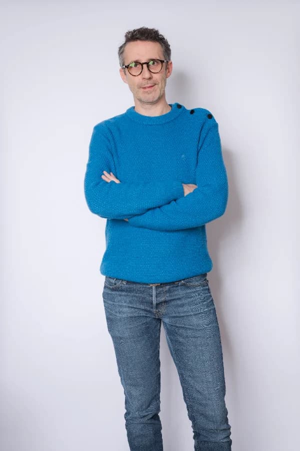 Photo de référence en studio : un homme à lunettes, pull tricot bleu et jean, bras croisés, debout devant un mur clair.