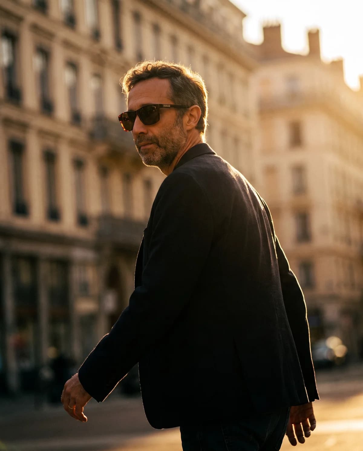 Founder portrait mid-stride, dark jacket, glancing back over the shoulder, raking backlight across a city square.