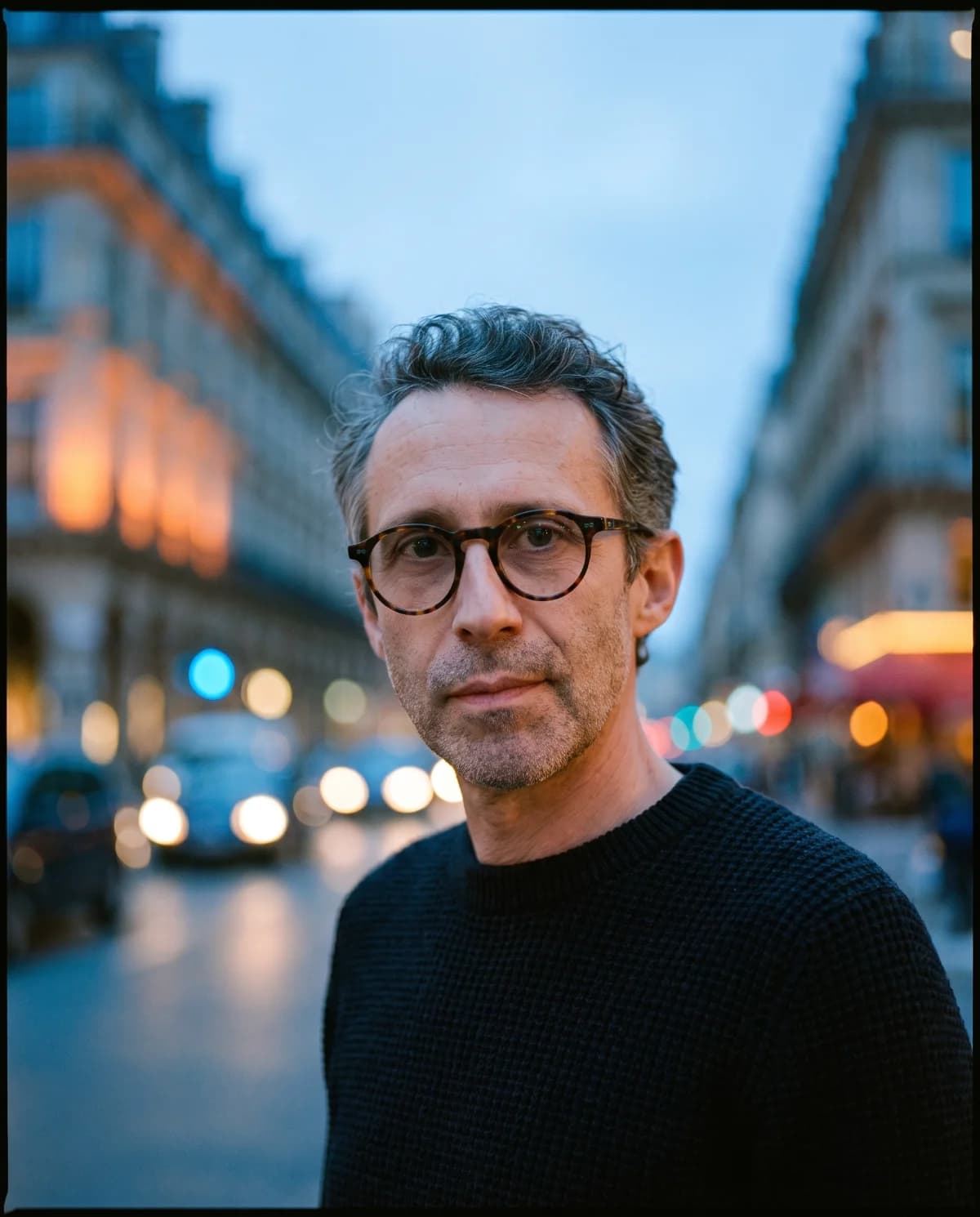 Close founder portrait in a dark knit and glasses at blue hour, street lights out of focus behind.
