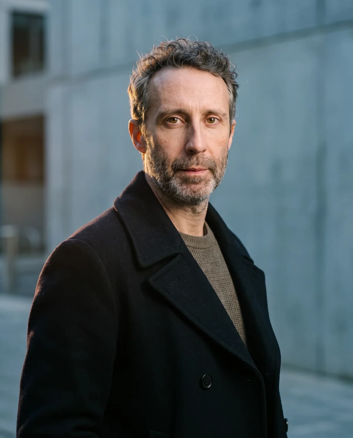 Founder portrait in a dark overcoat against a concrete wall, cold raking light, institutional register.